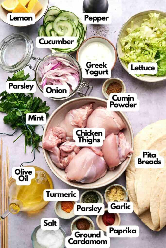 Ingredients for a dish are arranged on a surface, including chicken thighs, pita breads, spices, herbs, cucumber, onion, Greek yogurt, olive oil, lemon, lettuce, and mint.