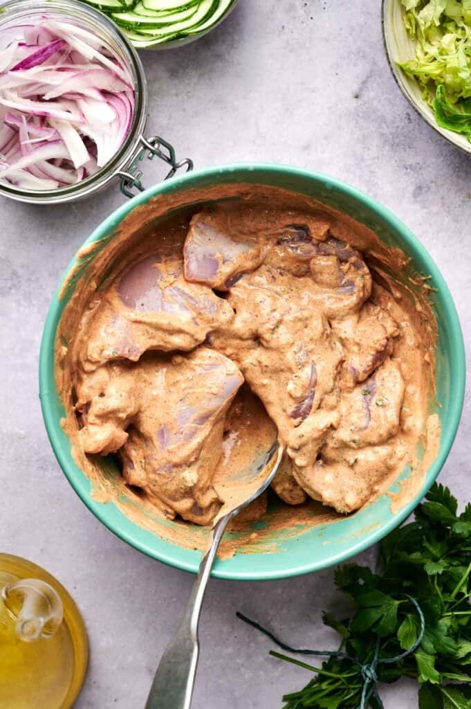 Bowl of raw chicken marinating in a creamy, spiced sauce with a spoon. Nearby are sliced onions, cucumber, lettuce, parsley, and a bottle of oil.