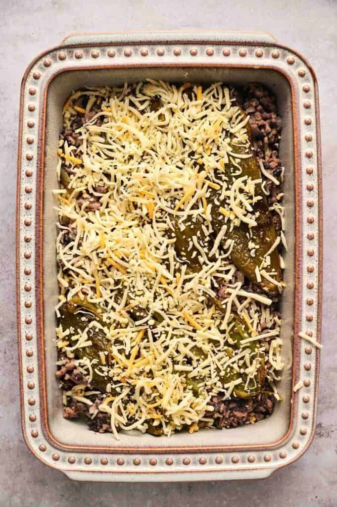 A baking dish layered with beef, sliced pickles, shredded cheese, and a hint of chili relleno casserole flavors, ready to be baked.