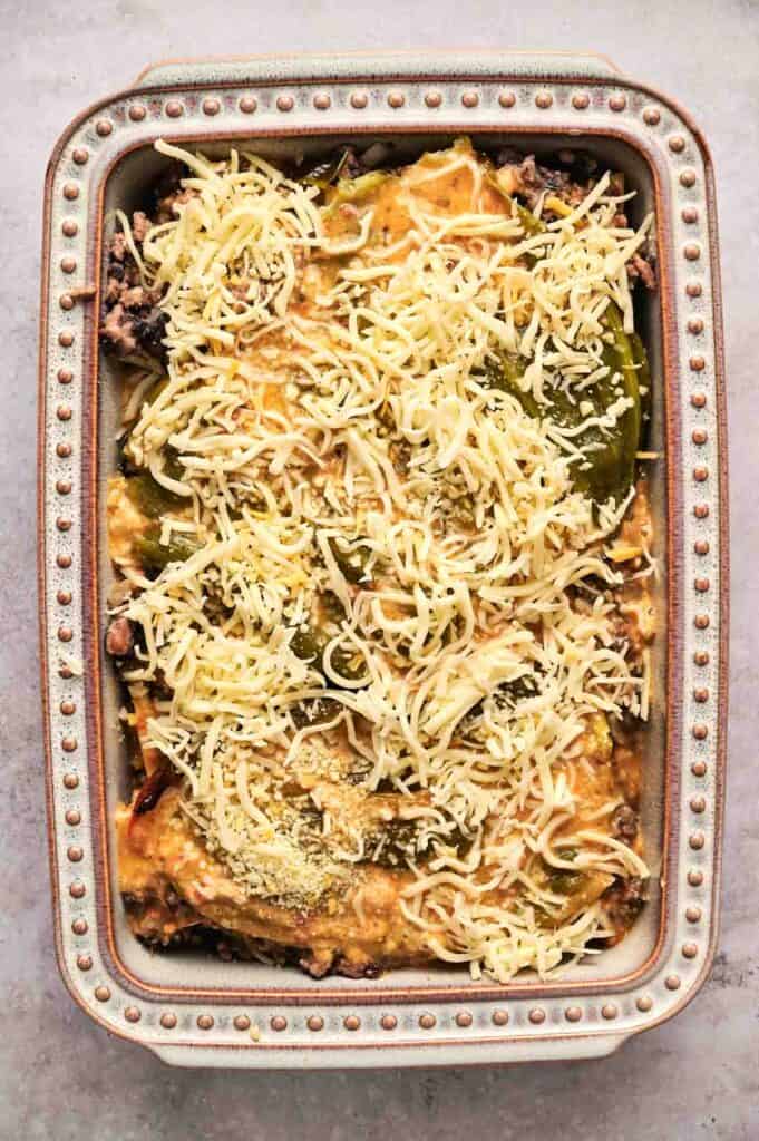 A rectangular baking dish filled with layers of pasta, meat, tomato sauce, and topped with shredded cheese, ready for baking, resembles a comforting chili relleno casserole.