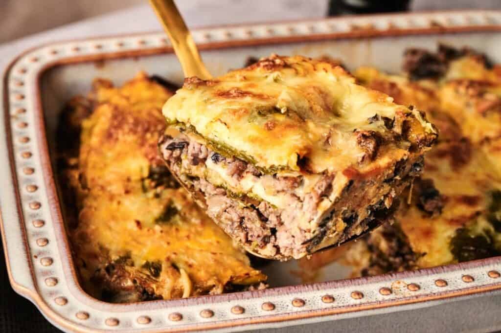 A slice of chili relleno casserole, with layers of meat, vegetables, and melted cheese, is being lifted from a baking dish.