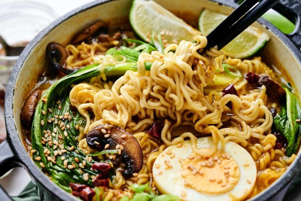 A bowl of Coconut Ramen with noodles lifted by chopsticks, topped with bok choy, mushrooms, a boiled egg, dried chilies, sesame seeds, and lime wedges.