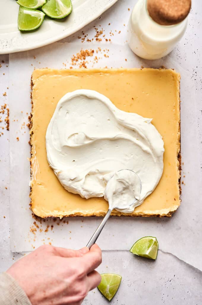 A hand spreads whipped cream over delicious Key Lime Bars on parchment paper, with lime wedges and a bottle nearby.