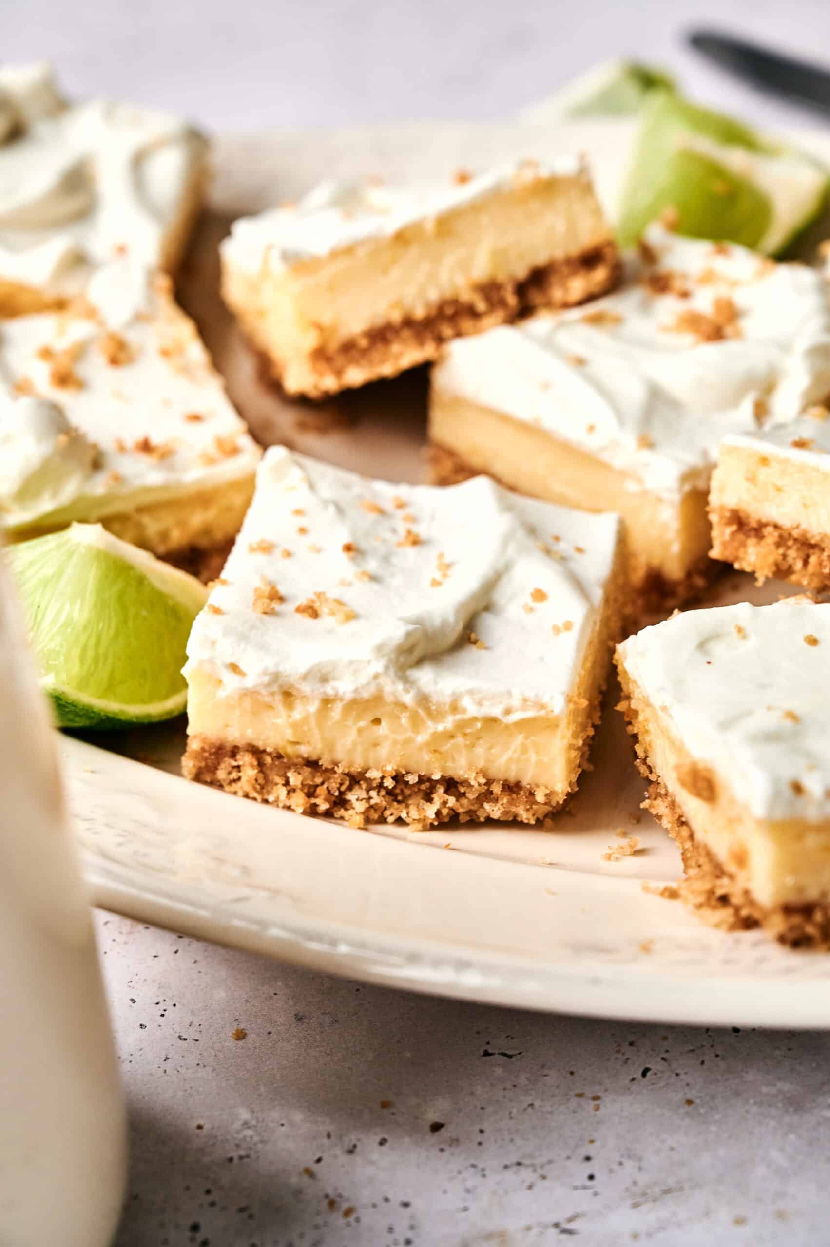 A plate of Key Lime Bars topped with whipped cream and graham cracker crumbs, with lime wedges in the background.