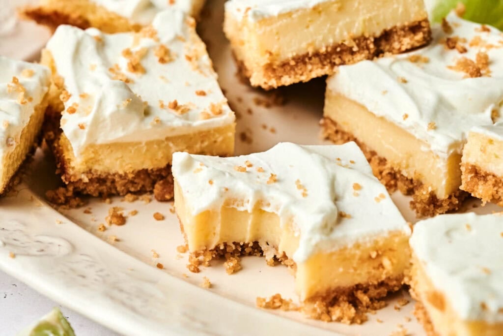 Several Key Lime Bars with a crumbly crust, creamy filling, and whipped topping are arranged on a plate; one has a bite taken out of it.