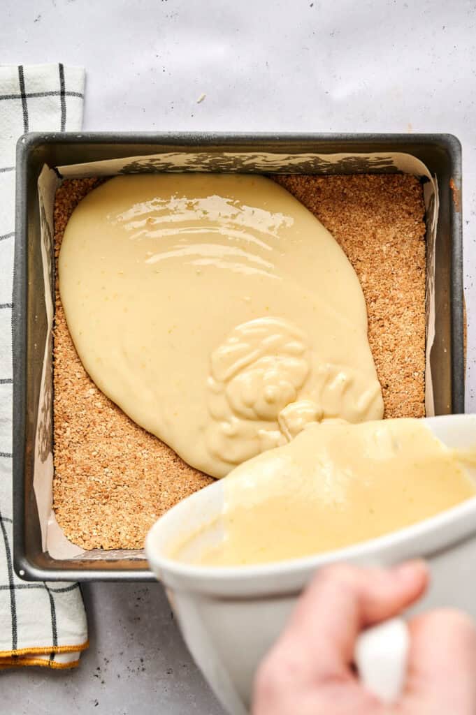 A hand pours creamy batter for Key Lime Bars from a bowl onto a graham cracker crust in a square baking pan lined with parchment paper.