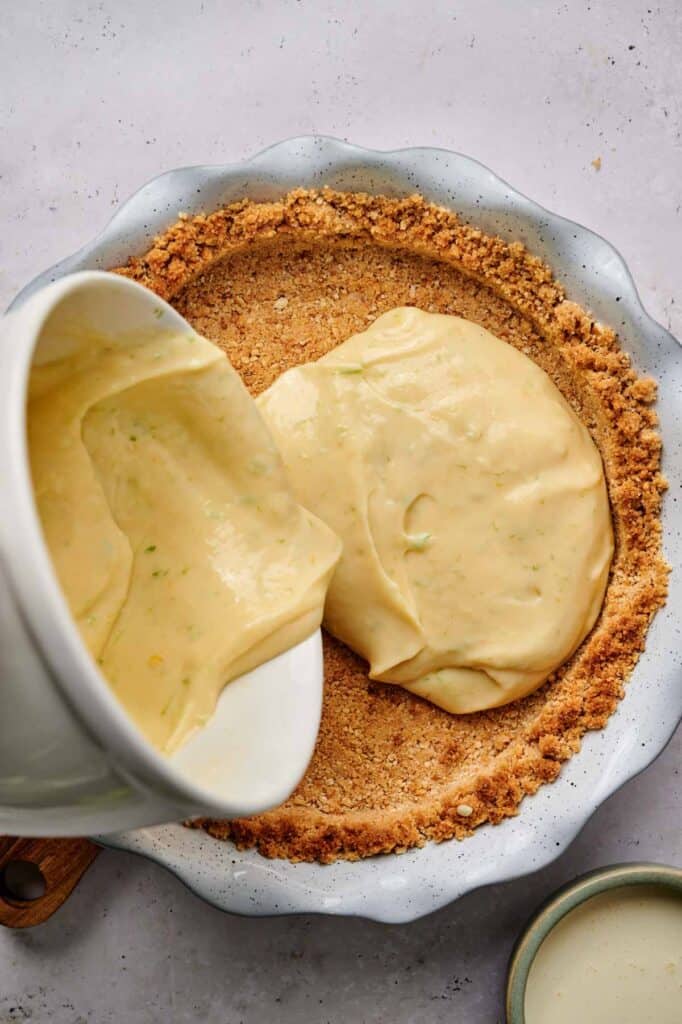 Pouring creamy key lime pie filling into a pie crust in a fluted dish on a gray surface.