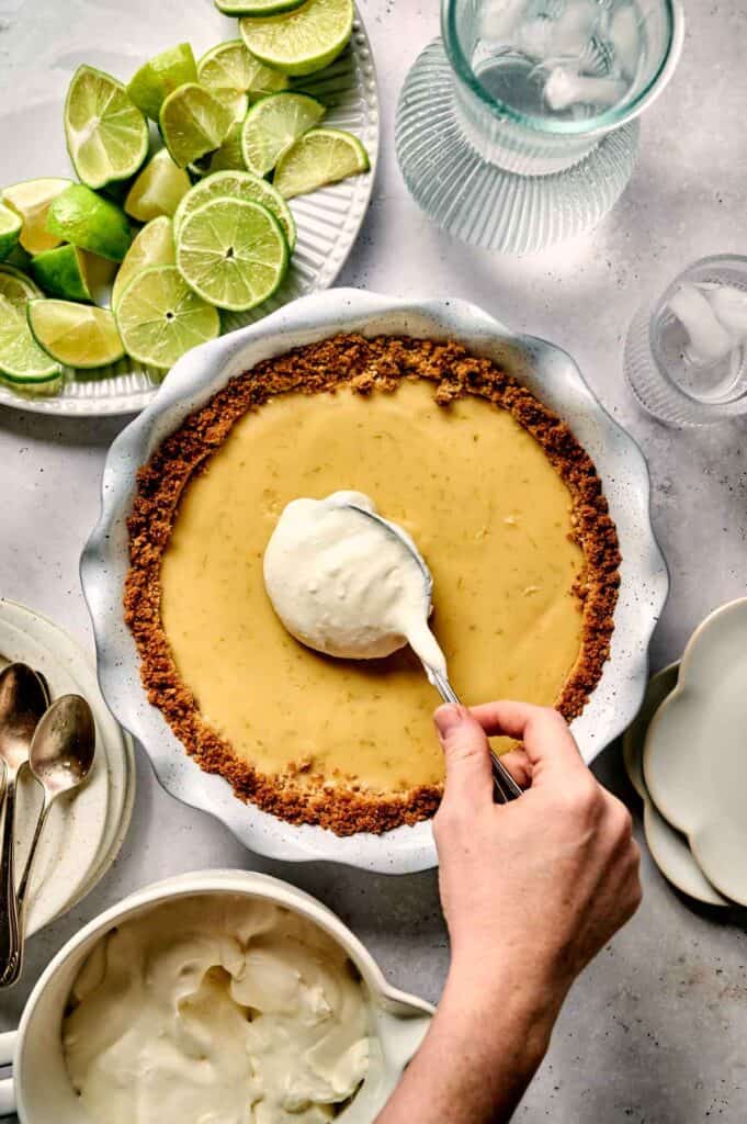 A hand skillfully spreads whipped cream over a tempting key lime pie in a white dish. Nearby, sliced limes add a fresh touch, while a glass of water and utensils await their time.