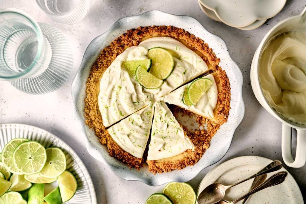 A sliced key lime pie with a graham cracker crust, topped with lime slices, sits on a white dish surrounded by plates, utensils, and fresh limes—capturing the zesty essence of classic key lime pie.