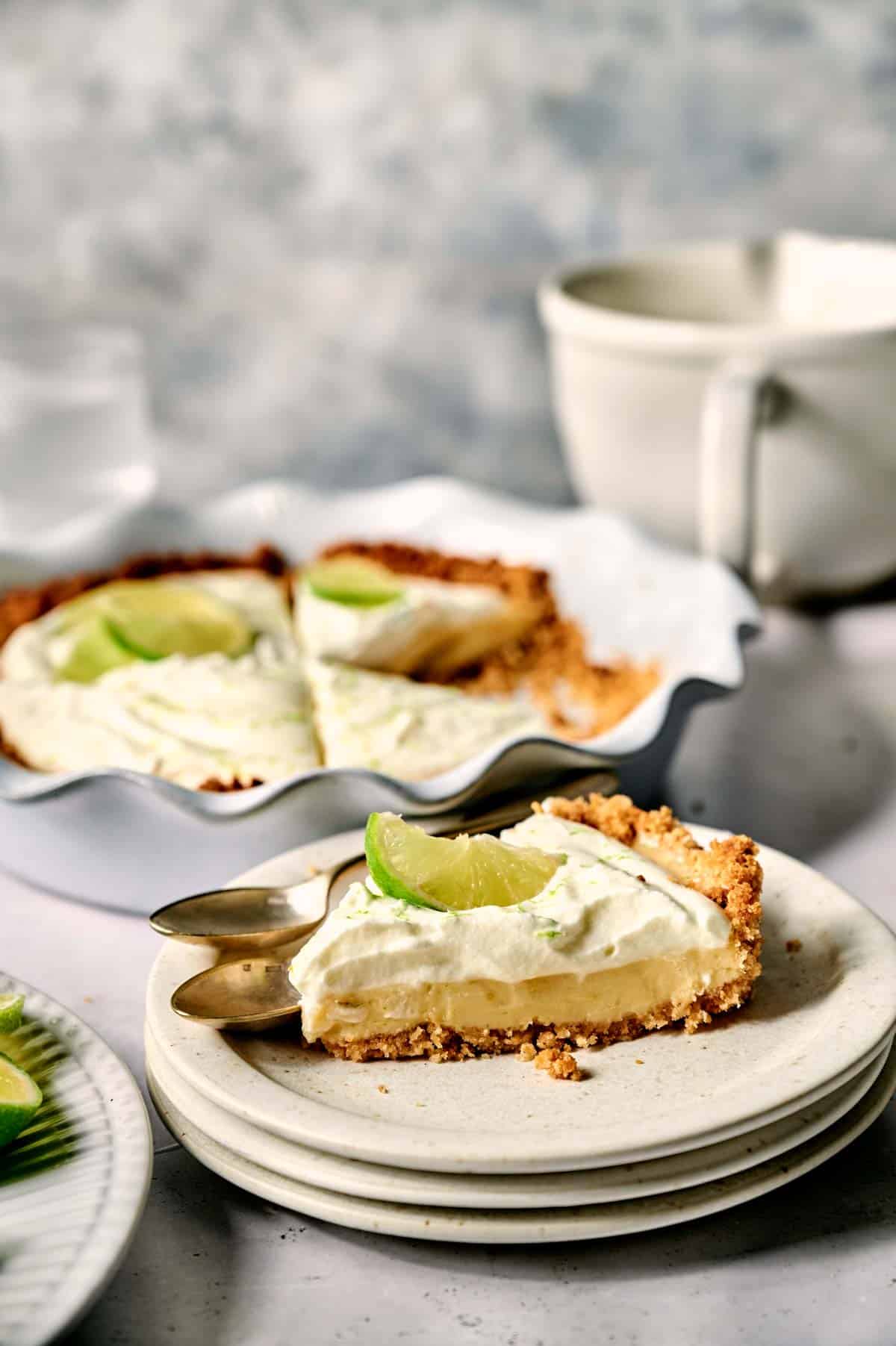 A slice of key lime pie with a buttery graham cracker crust and a fresh lime garnish rests elegantly on stacked plates, while the remaining key lime pie beckons from the dish in the background.