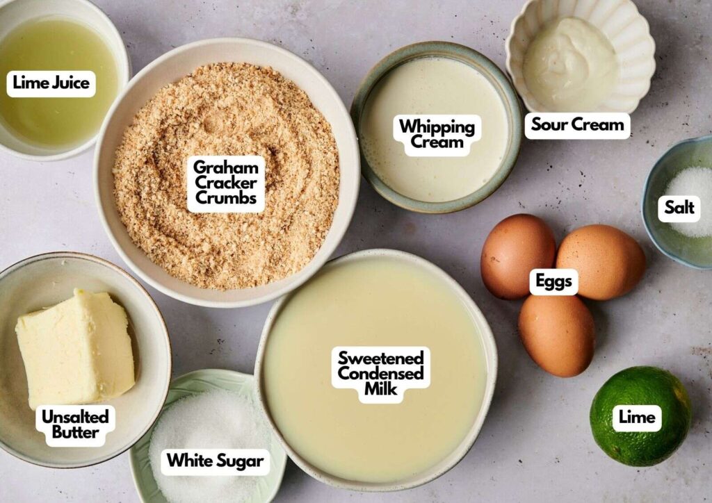 Bowls containing labeled ingredients for a key lime pie: lime juice, graham cracker crumbs, whipping cream, sour cream, salt, eggs, sweetened condensed milk, unsalted butter, white sugar, and a lime.