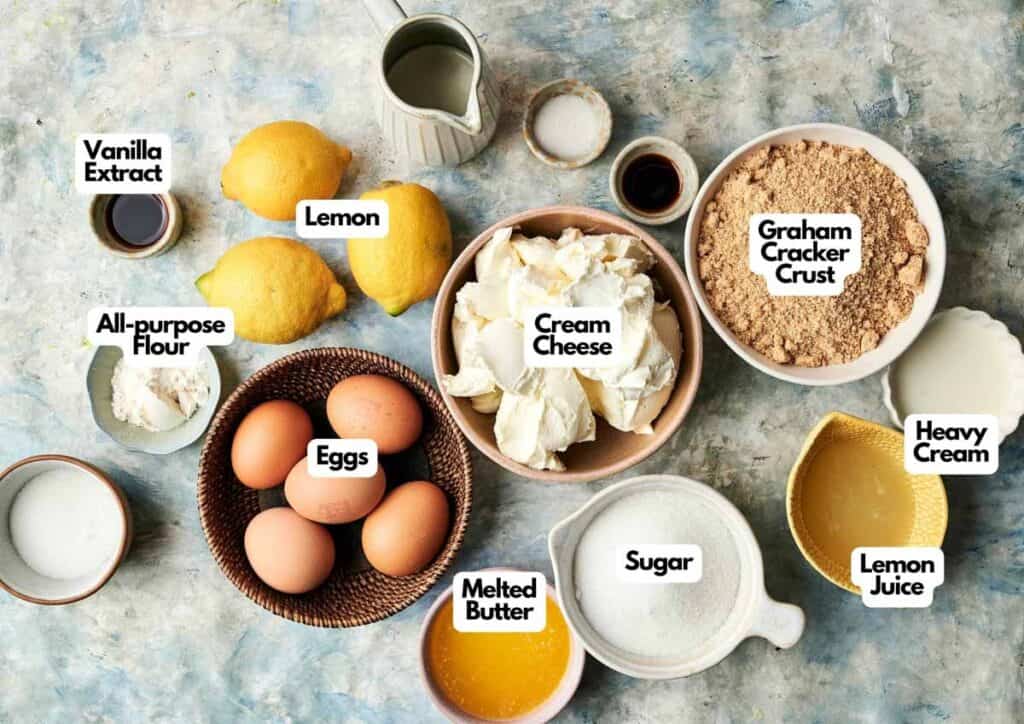 Top view of labeled lemon cheesecake ingredients, including cream cheese, eggs, sugar, vanilla extract, heavy cream, graham cracker crust, melted butter, flour, lemons, and lemon juice.