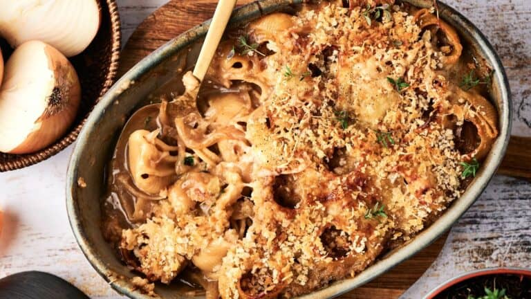 Oval baking dish filled with baked onion casserole topped with golden breadcrumbs and herbs; a wooden spoon rests in the dish. Sliced onions are visible on the side.