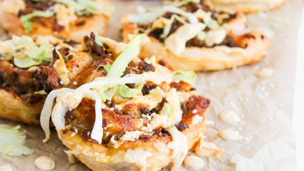 Small puff pastry rounds topped with ground meat, shredded lettuce, melted cheese, sesame seeds, and sauce, arranged on parchment paper.