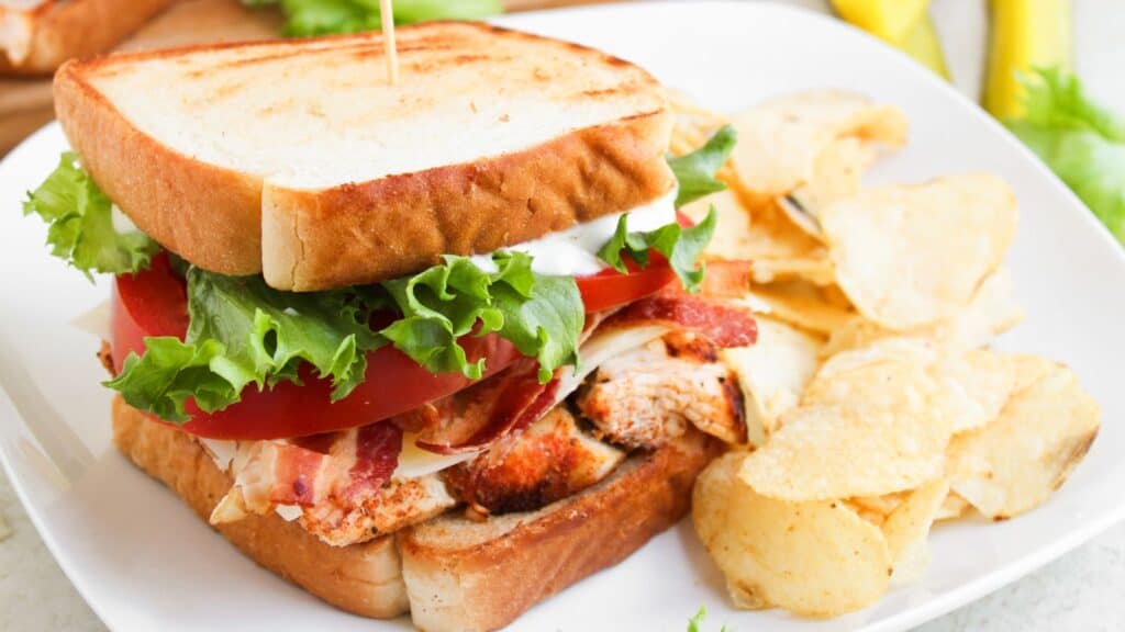 A turkey club sandwich with lettuce, tomato, and bacon on toasted bread is served on a white plate beside a pile of potato chips.