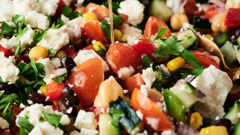 A close-up of a colorful salad with tomatoes, feta cheese, chickpeas, black beans, corn, red onion, parsley, and a drizzle of dressing.