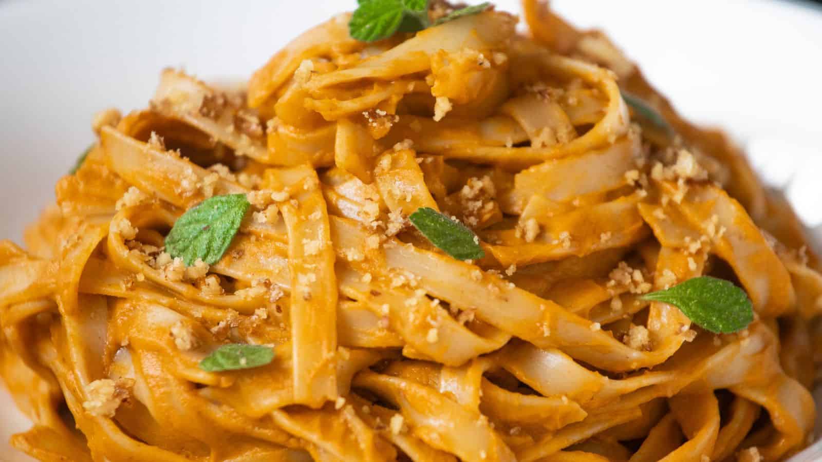 A close-up of tagliatelle pasta coated in a creamy orange sauce, topped with chopped nuts and fresh green herbs.