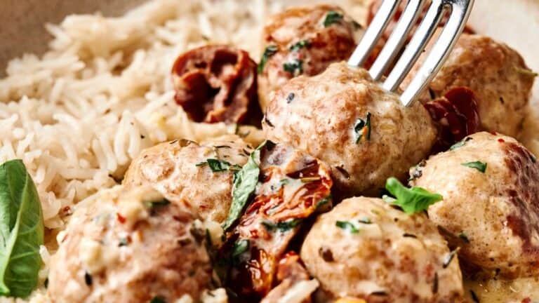 A close-up of meatballs with creamy sauce, herbs, and sun-dried tomatoes, served alongside a portion of white rice.