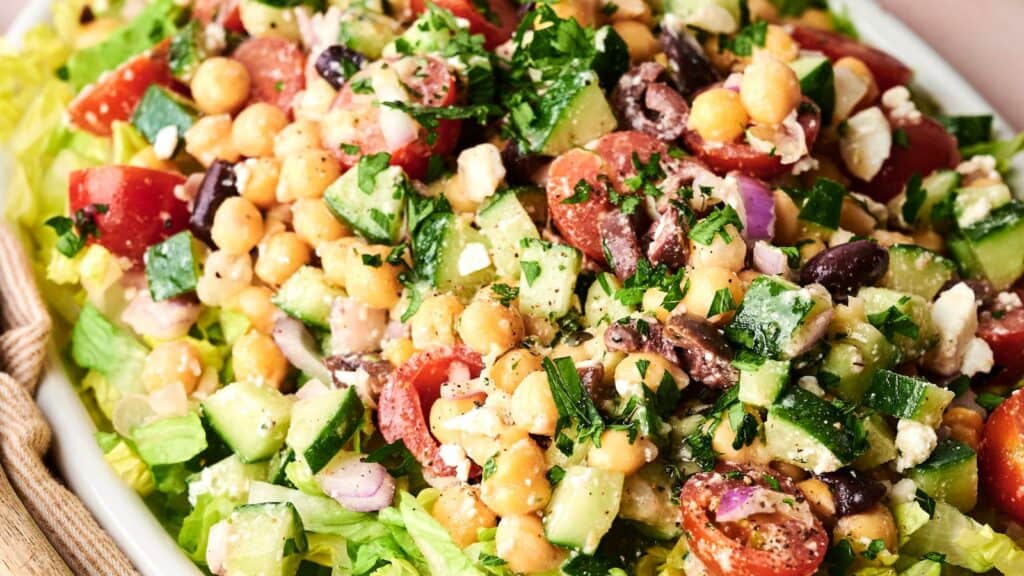 A close-up of a Mediterranean salad with chopped cucumbers, tomatoes, chickpeas, red onion, feta cheese, olives, and parsley on a bed of lettuce.
