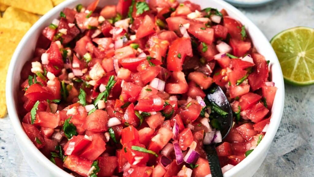 A white bowl filled with freshly made pico de gallo, featuring chopped tomatoes, onions, cilantro, and jalapeños, with a spoon inside. A sliced lime and tortilla chips are nearby.