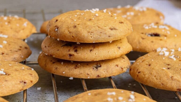 Three chocolate chip cookies stacked on a cooling rack, surrounded by more cookies and sprinkled with coarse salt.