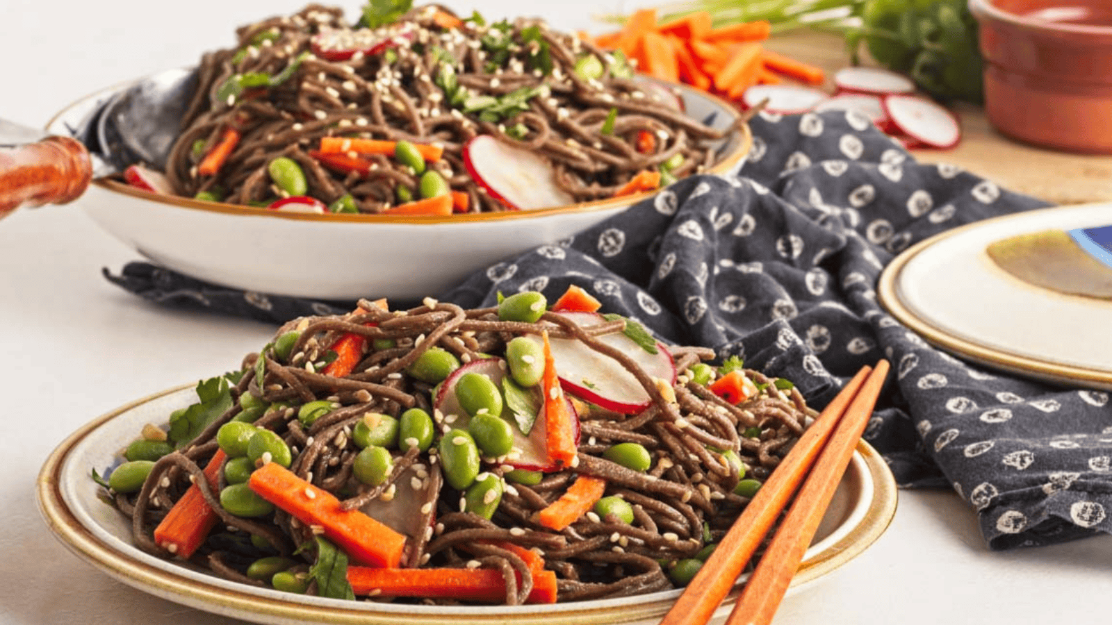 Two bowls of soba noodle salad with carrots, edamame, and radish slices, placed on a patterned cloth with wooden chopsticks beside them.