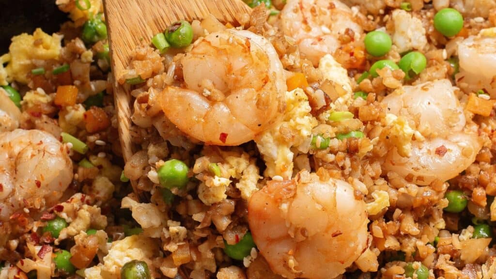 Close-up of shrimp fried rice with peas, scrambled egg, carrots, and green onions being stirred with a wooden spatula.
