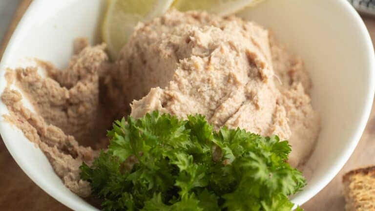 A white bowl filled with a creamy tuna dip, garnished with fresh parsley and lemon slices.