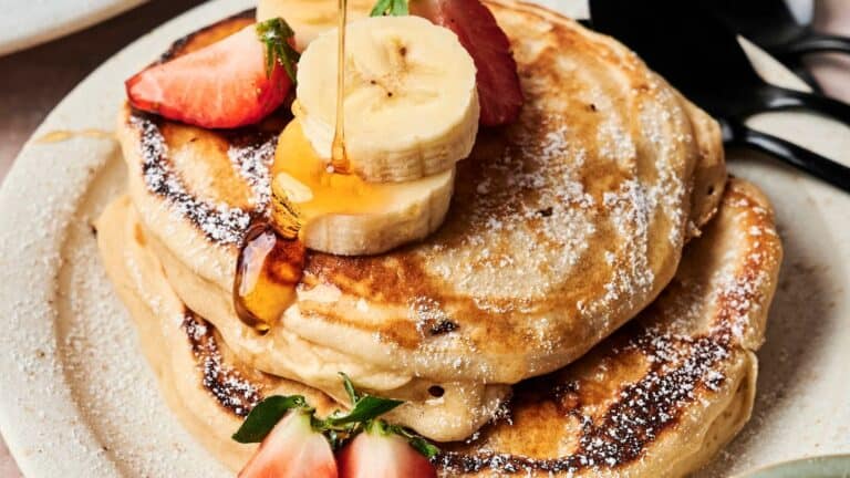 A stack of pancakes topped with banana slices, strawberries, and a drizzle of syrup on a white plate.