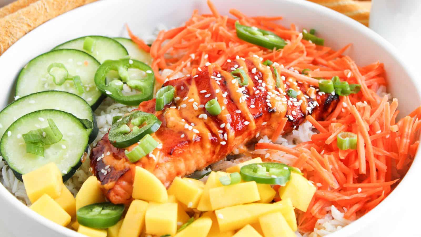 A bowl with grilled salmon on rice, topped with sesame seeds, surrounded by sliced cucumbers, shredded carrots, jalapeños, mango cubes, and chopped green onions.