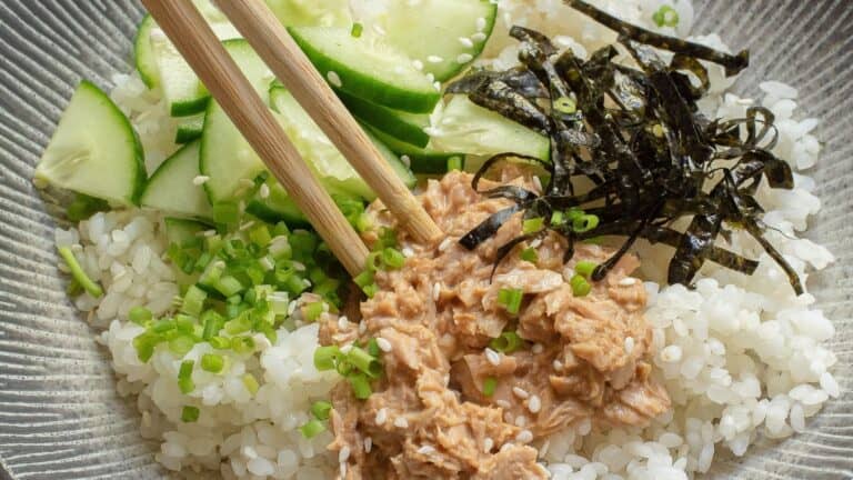 A bowl of white rice topped with chopped cucumber, shredded seaweed, tuna, and chopped green onions, with a pair of wooden chopsticks placed on top.
