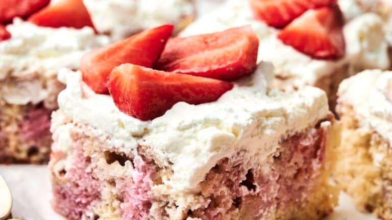 Close-up of a cake slice topped with whipped cream and strawberry pieces. The cake has a marbled pink and white appearance.