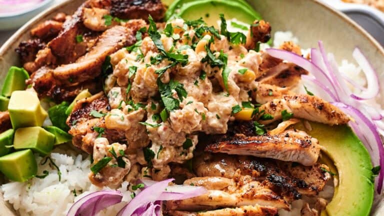 A dish with grilled chicken slices, creamy sauce, avocado slices, chopped cilantro, rice, and red onion slivers.