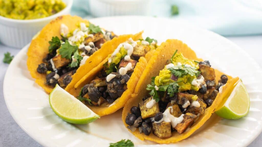 Three corn tortilla tacos filled with black beans, diced vegetables, and topped with creamy sauce and herbs, served with lime wedges on a white plate.