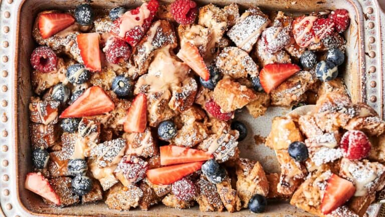 Baked French toast casserole topped with strawberries, raspberries, blueberries, and powdered sugar in a rectangular dish.