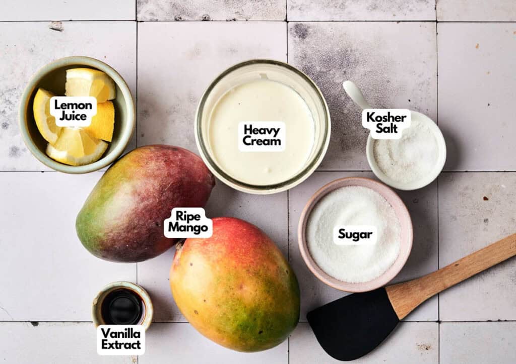 Ingredients for mango ice cream are laid out on a tiled surface: lemon juice, heavy cream, kosher salt, sugar, ripe mangoes, vanilla extract, and a black spatula.