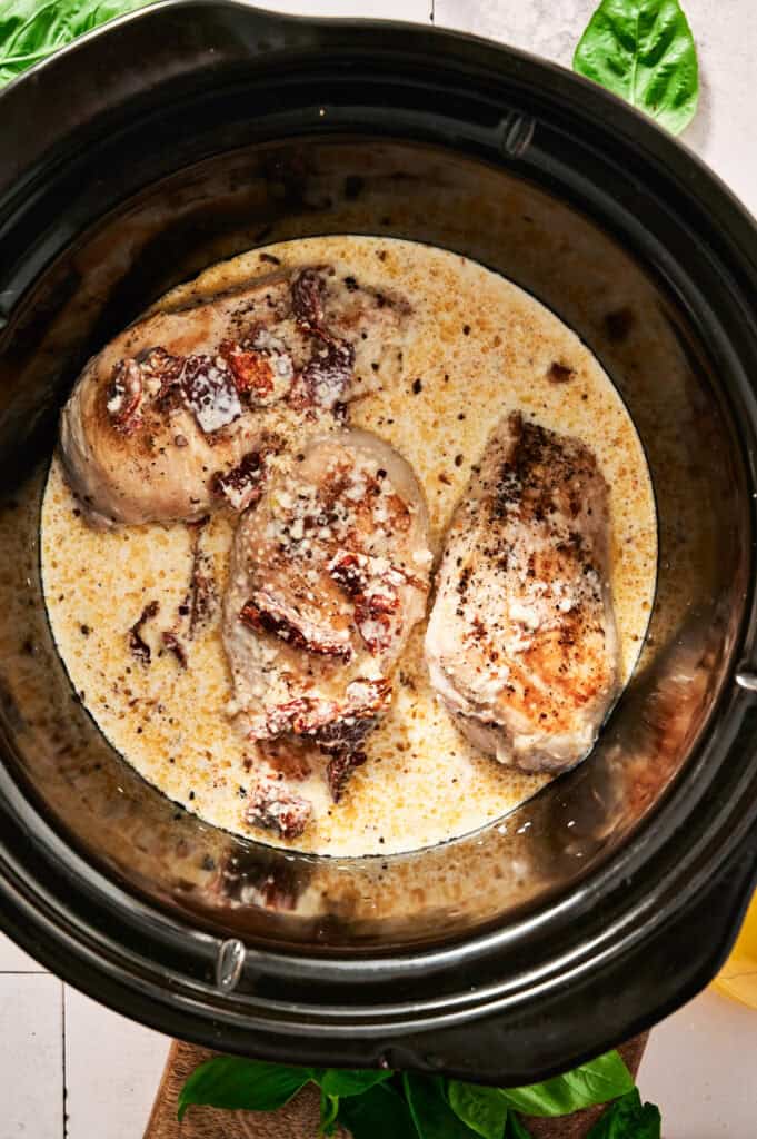 Crock Pot Marry Me Chicken: Three tender cooked chicken breasts in a creamy sauce, adorned with sun-dried tomatoes, nestled perfectly inside a slow cooker. Fresh basil leaves add the final touch of flavor and aroma.