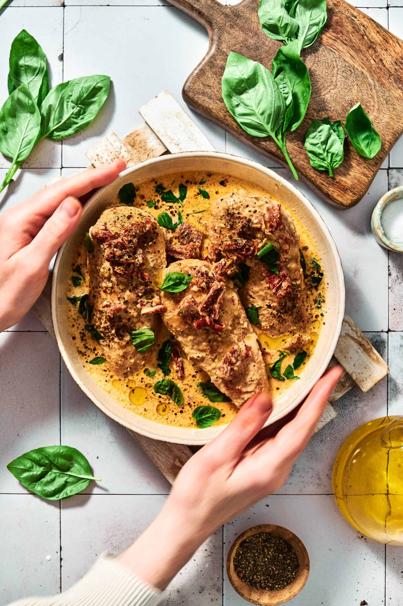 Hands holding a bowl of creamy Crock Pot Marry Me Chicken garnished with herbs. Basil leaves and a wooden board with more leaves are nearby. A small dish of black pepper and olive oil are also visible, enhancing the inviting aroma.