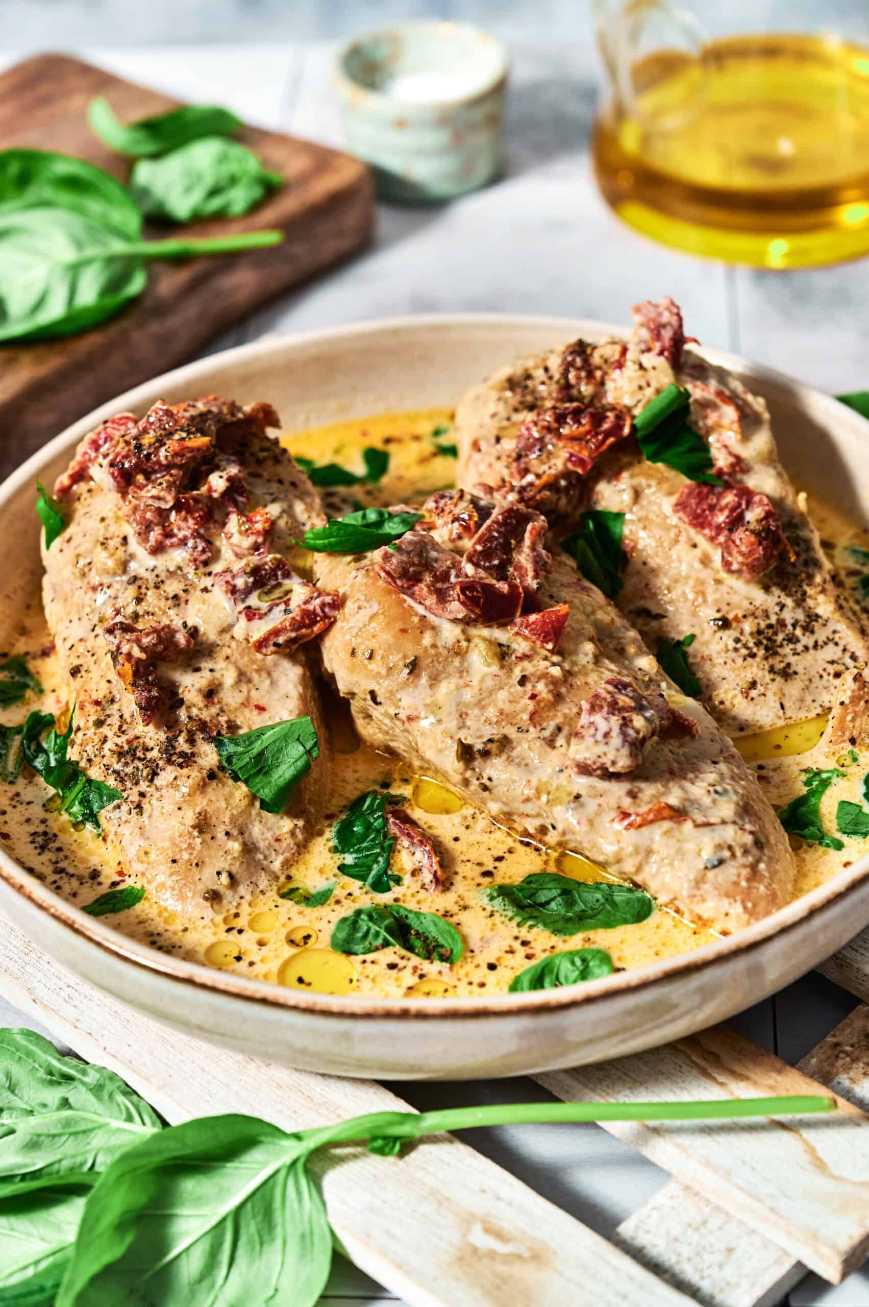 Chicken breasts in a creamy sauce with sun-dried tomatoes and fresh basil, reminiscent of Crock Pot Marry Me Chicken, served in a beige dish; olive oil and additional basil leaves in the background.