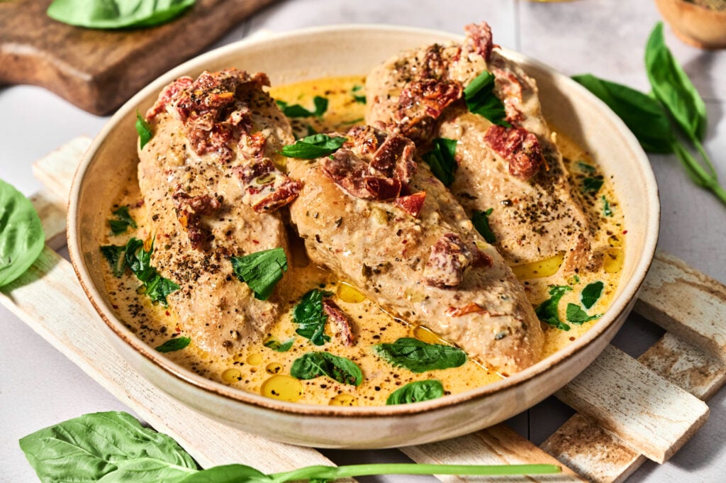 Three chicken breasts in a creamy sauce garnished with sun-dried tomatoes and fresh basil leaves, served in a round bowl—an irresistible Crock Pot Marry Me Chicken dish perfect for any occasion.