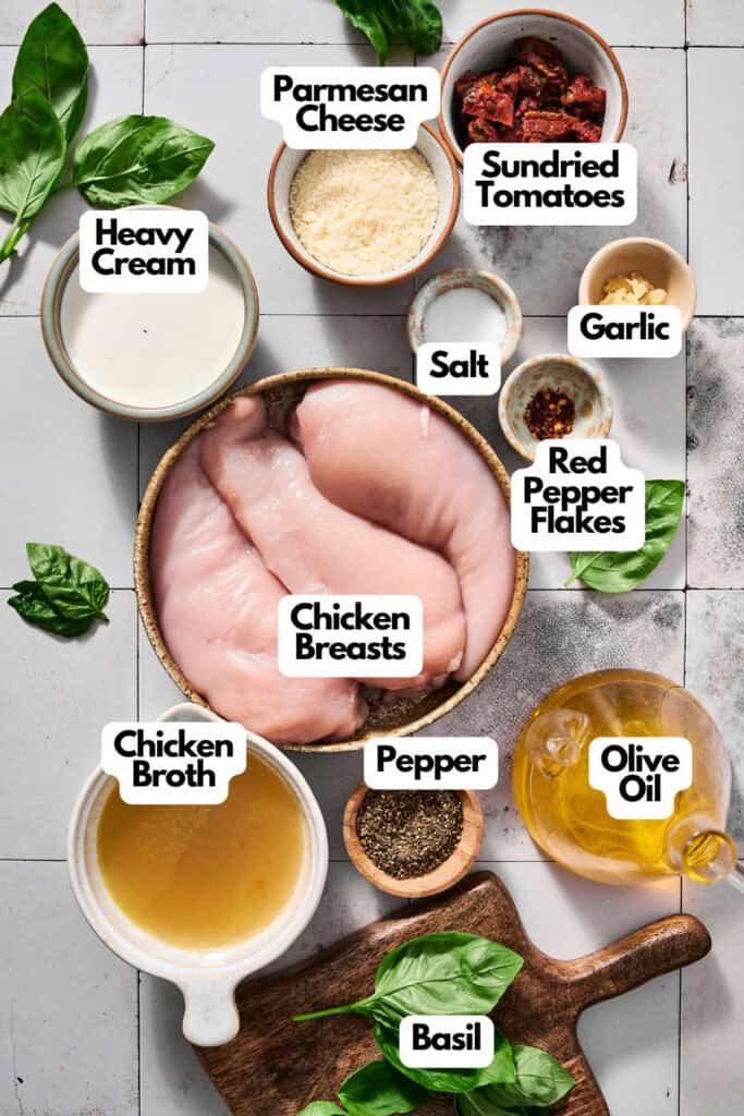 Ingredients laid out on a table for Crock Pot Marry Me Chicken, featuring chicken breasts, heavy cream, Parmesan cheese, sundried tomatoes, garlic, red pepper flakes, salt, pepper, chicken broth, olive oil, and basil.