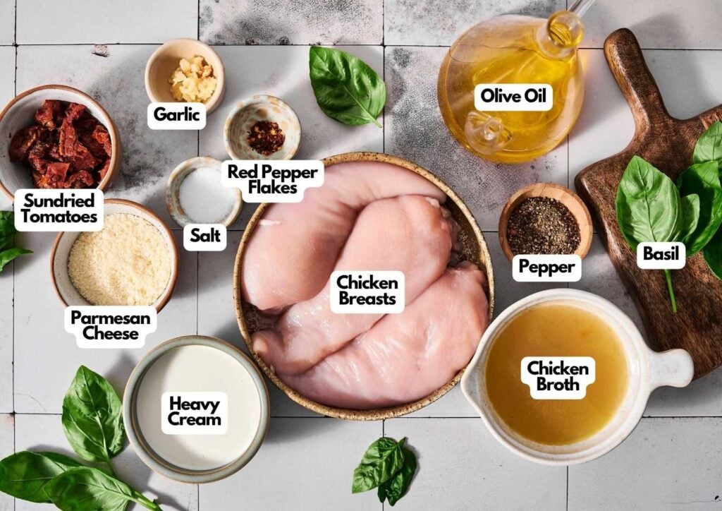 A flat lay of ingredients for Crock Pot Marry Me Chicken, featuring chicken breasts, heavy cream, parmesan, sundried tomatoes, garlic, red pepper flakes, salt, pepper, olive oil, chicken broth, and basil.