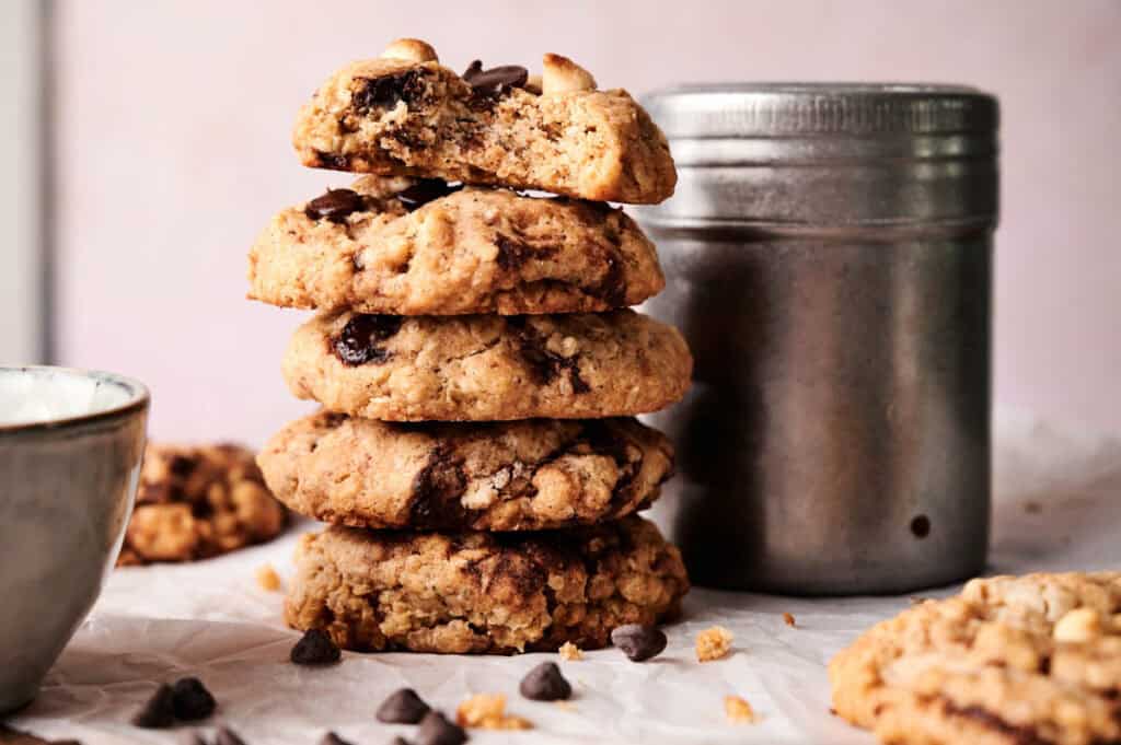 A stack of four chocolate chip cookies on parchment paper, with one partially eaten on top, sits near a metal container and a ceramic cup—perfect companions for an indulgent ice cream cake dessert.