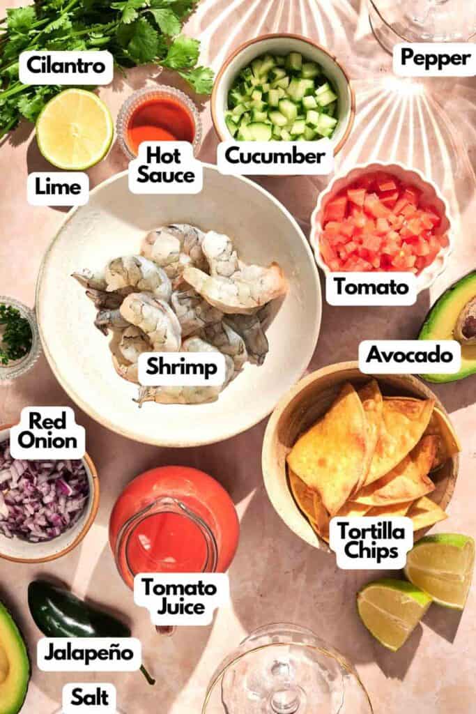 An assortment for a Mexican Shrimp Cocktail awaits, with raw shrimp, diced vegetables, and tortilla chips laid out alongside zesty condiments like lime, hot sauce, and pepper on the table.