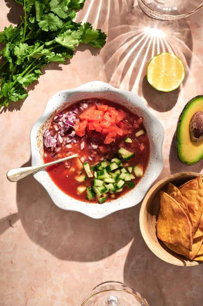 A bowl of refreshing Mexican Shrimp Cocktail-inspired tomato soup, topped with diced tomatoes, cucumbers, and onions. Accompanied by cilantro, a lime half, creamy avocado slices, and a side of crunchy tortilla chips for the perfect touch.