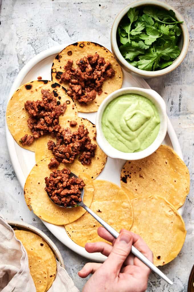 A hand spoons cooked ground meat onto corn tortillas on a platter, creating delicious mini tacos, with bowls of green sauce and fresh cilantro beside them.