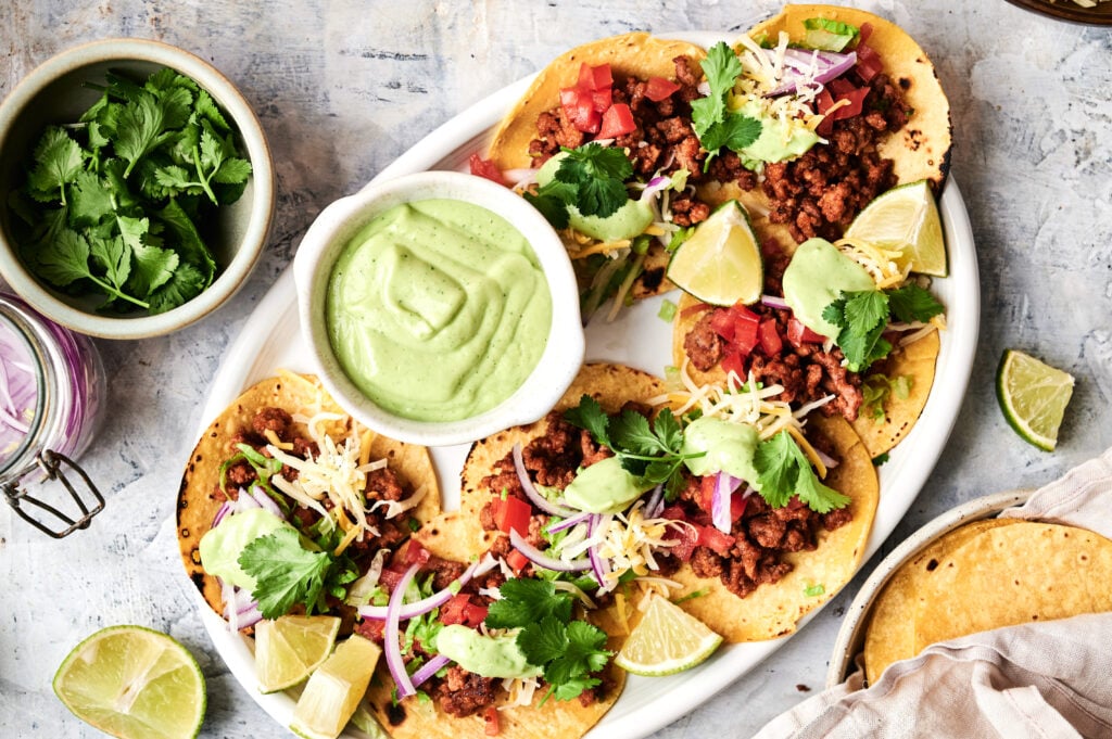 A plate of mini tacos with ground meat, shredded cheese, tomato, onion, cilantro, and lime wedges, served with a bowl of creamy green sauce and a side of fresh cilantro.