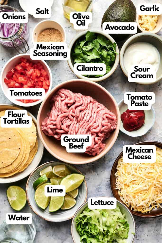Ingredients for making mini tacos are arranged on a table, including ground beef, tortillas, avocado, cheese, cilantro, lettuce, sour cream, lime, and various seasonings.