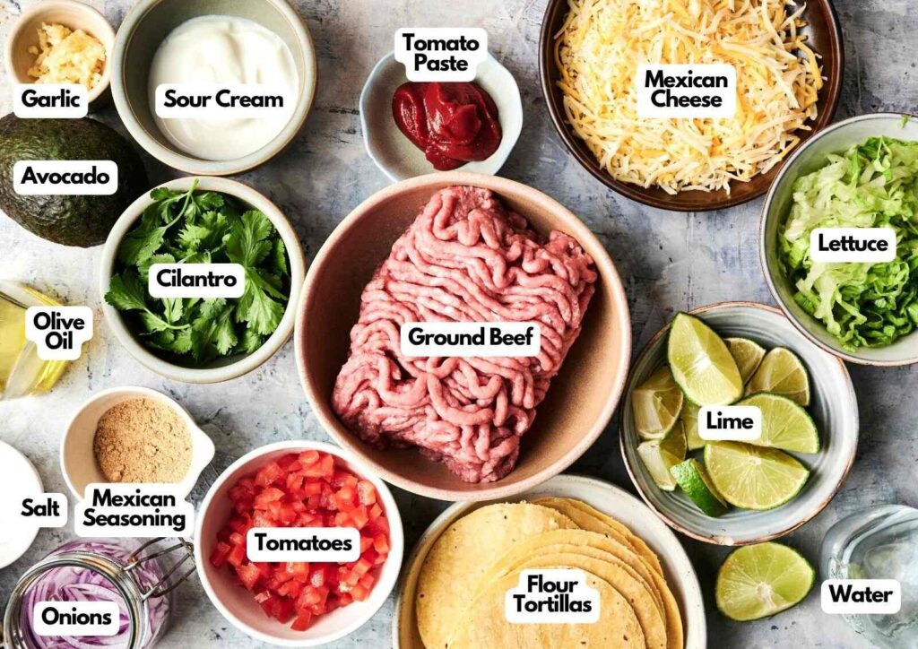 Various taco ingredients arranged in bowls and plates—including ground beef, cheese, tomatoes, lettuce, cilantro, lime, tortillas, avocado, sour cream and seasonings—perfect for assembling your own delicious mini tacos at home.