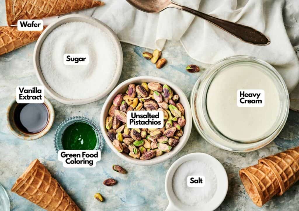 Top-down view of Pistachio Ice Cream ingredients, including sugar, heavy cream, unsalted pistachios, salt, green food coloring, vanilla extract, and wafer cones arranged on a marble surface.
